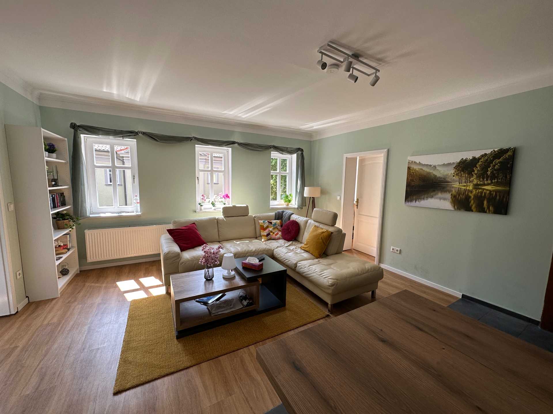 Weiße Wohnlandschaft vor drei Fenstern, gelber Teppich mit kleinem Tisch, weißes Regal, Trennungswohnung